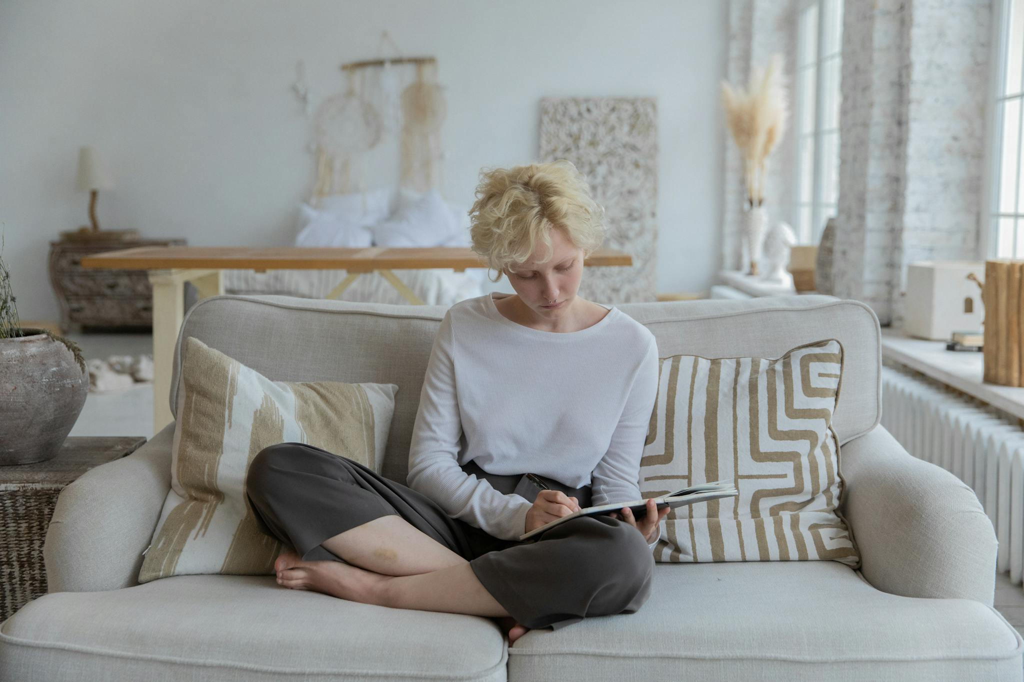 Full body of young barefooted pensive lady with curly blond hair in casual clothes sitting on soft couch and writing thoughts in notebook during remote work at home