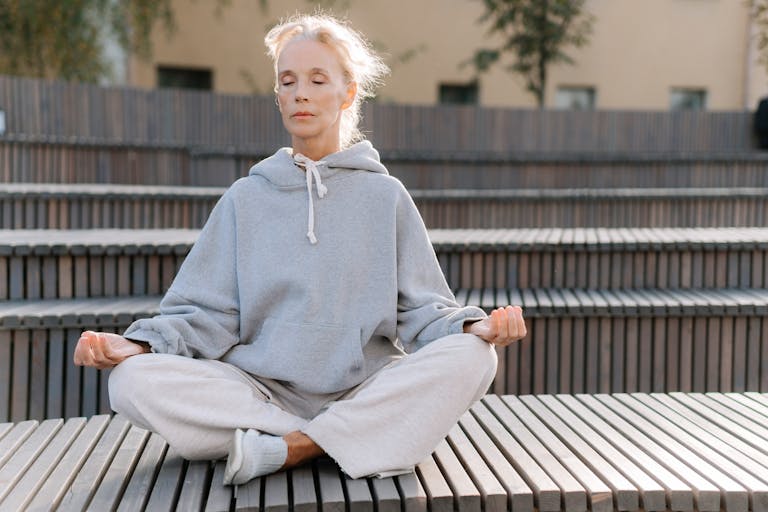 Elderly woman in gray hoodie meditating outdoors, promoting fitness and relaxation.